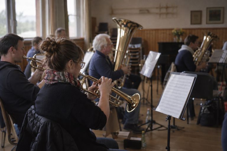 Probe einer Blaskapelle im Probenraum: Vorn spielt eine Trompeterin mit Brille und Schal vom Notenständer, neben ihr ein Trompeter, dahinter weitere Musizierende an Tuba und anderen großen Blechblasinstrumenten.