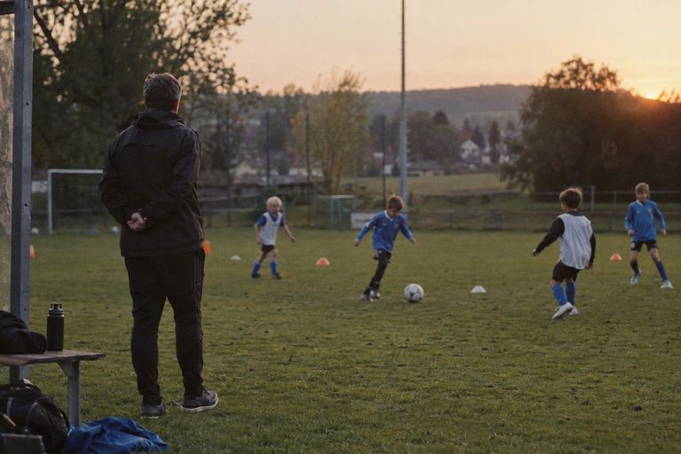 Jugendtraining auf dem Sportplatz in der Abendsonne: Ein Trainer steht mit verschränkten Händen am Spielfeldrand und schaut vier Kindern in Trikots und Leibchen beim Passspiel zwischen Hütchen zu.