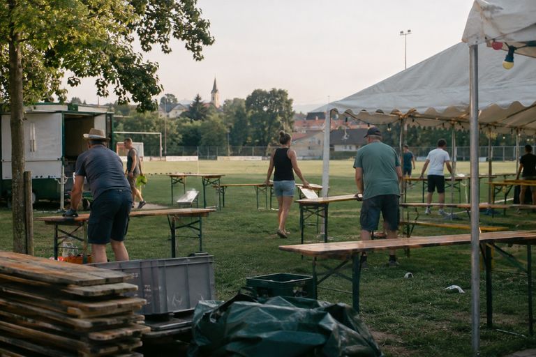 Ehrenamtliche bauen auf einem Sportplatz ein Vereinsfest auf: Sie tragen Bierbänke und Tische über den Rasen und stellen sie neben einem Festzelt mit Lichterkette auf. Vorn liegen gestapelte Holzplatten und Kisten, im Hintergrund stehen ein Fußballtor und ein Kirchturm.