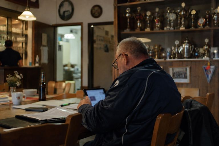 Ein Vorstandsmitglied mit Brille und Trainingsjacke sitzt im Vereinsheim am Tisch und schaut auf ein Tablet, daneben liegen Unterlagen. Im Hintergrund stehen Pokale im Regal, hinten an der Theke steht eine weitere Person.